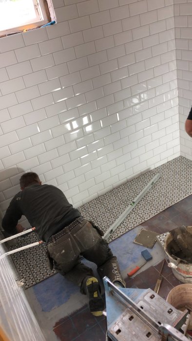Arbetare lägger kakel på tvättstugegolv med vita väggkakel i bakgrunden.
