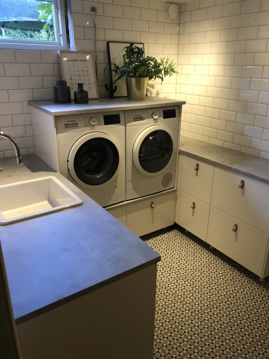 Renoverad tvättstuga med vita skåp, tvättmaskin, torktumlare, subway-kakel och mönstrat golv.