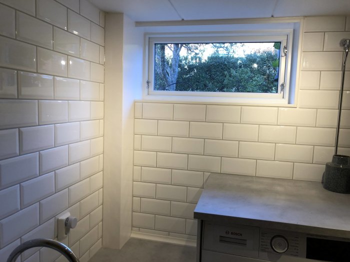 Nyrenoverad tvättstuga med vita kakelväggar, ett fönster och en Bosch tvättmaskin.