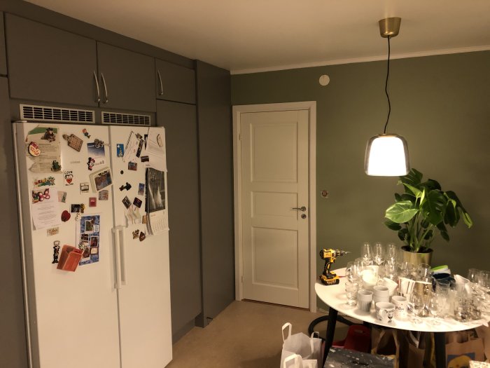 Uppfräschade köksskåp i grått, ett kylskåp täckt med magneter och lappar, en hängande lampa och en bord med glas.
