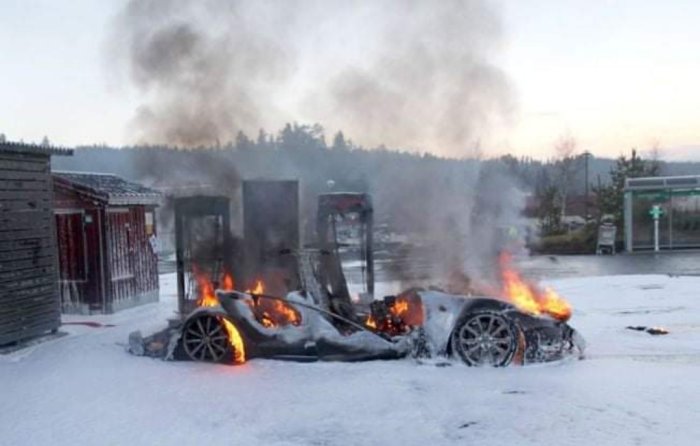 Elbil i brand vid laddstation med rök och flammor, illustrerar risk med ellbilsladdning.