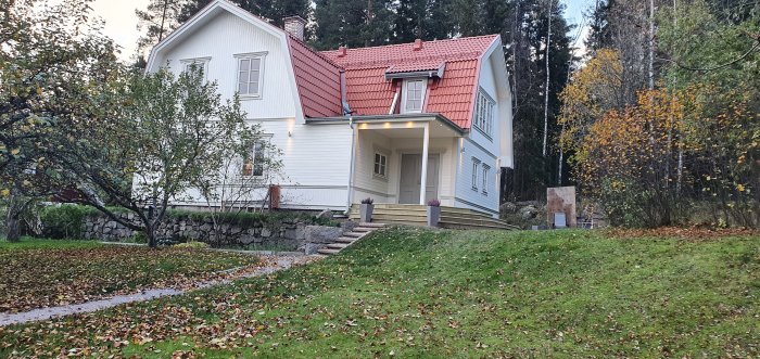 Renoverat hus från 1926 med ny balkong ovanför entrén, vit fasad och rött tegeltak omgiven av grönska.