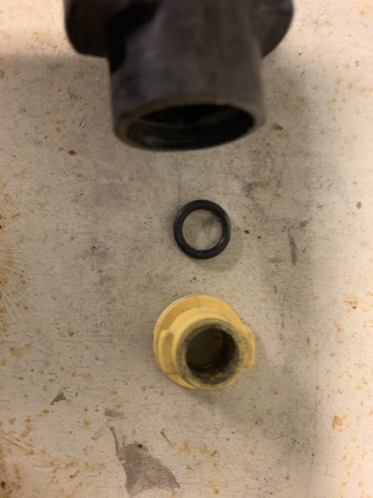 Närbild av en demonterad rördel, en svart O-ring och en gul koppling, på en smutsig yta.