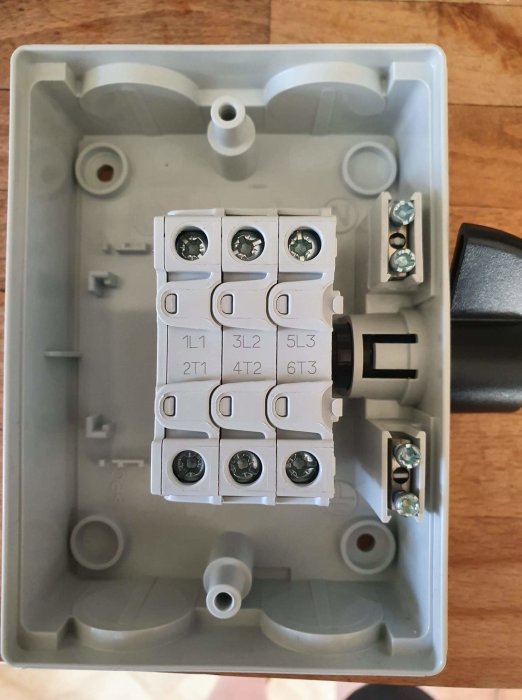 Elektrisk huvudbrytare i en grå kopplingslåda med etiketterade terminaler och en kabelanslutning.