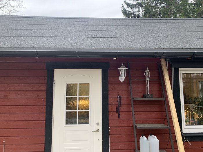 Sadeltak med takpapp och hängrännor på en rödmålad byggnad med vit dörr och fönster.