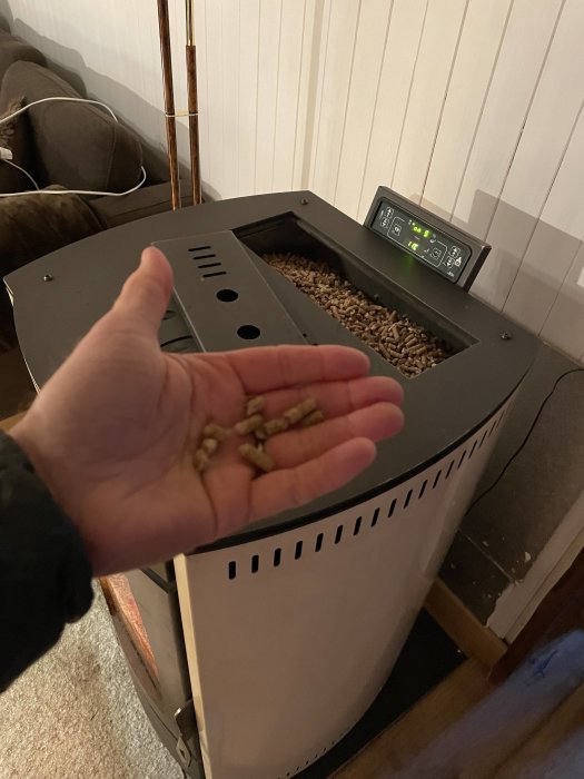 Hand som håller träpellets framför en öppen pelletskamin med digitala kontroller.