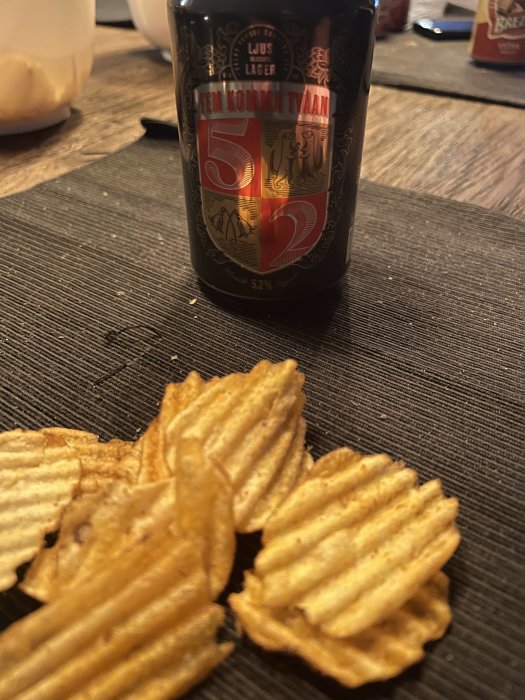 Ölflaska och potatischips i förgrunden på ett bord med suddig bakgrund.