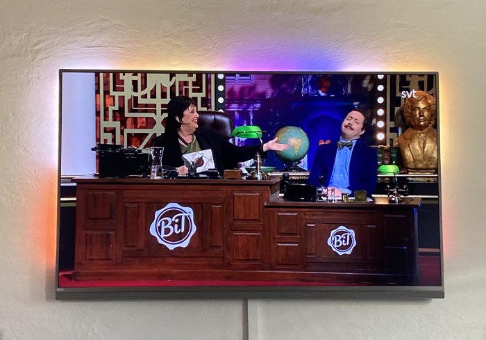 Två personer skrattar bakom ett skrivbord med diverse föremål på en TV-skärm som visar SVT-program.