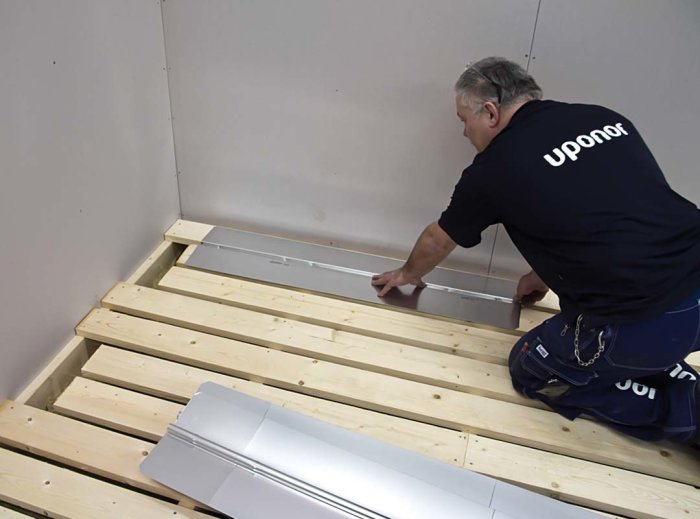 Person installerar golvvärmeplåtar mellan träbjälkar på ett spåntat golv.