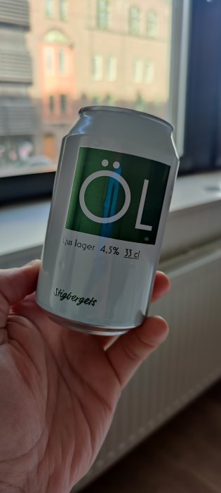 Hand håller en burk ljus lageröl framför ett fönster i ett hotellrum.