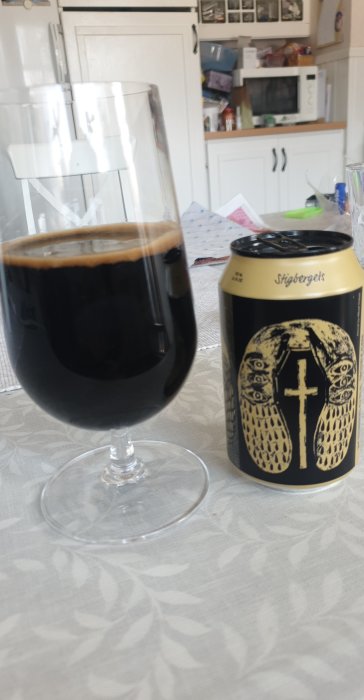 Ett glas med mörkt öl bredvid en ölburk med etikett, står på ett bord med vit duk.