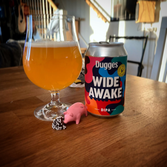 Ölglas fyllt med guldfärgad dryck bredvid en färgglad burk märkt 'Dugges WIDE AWAKE DIPA' och ett litet rosa grissparbössa.