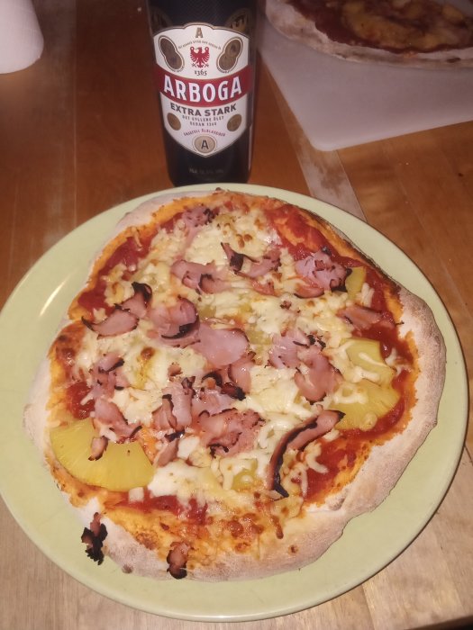 Hemlagad panpizza på tallrik bredvid en flaska öl, med skinka och ananas på.