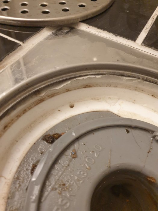 Närbild på en felaktigt skuren brunnsmanchett i en golvbrunn, omgiven av klinker och smuts.