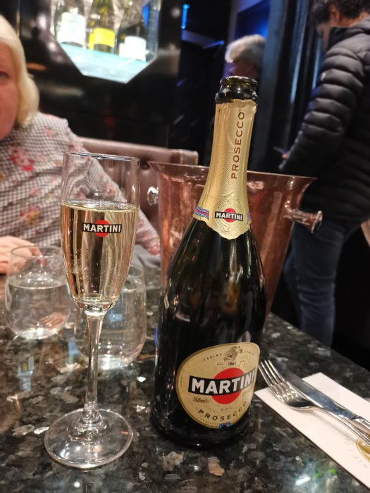 Flaska och glas med Martini Prosecco på bord framför person i restaurangmiljö.