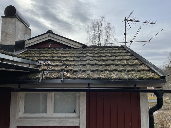 Mossbelagt tak med troligtvis eternitplattor, antenner ovanpå och villa i bakgrunden.