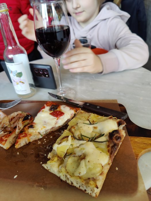 Potatispizza med rosmarin på en skärbräda, ett glas rödvin och en delvis suddig person i bakgrunden.