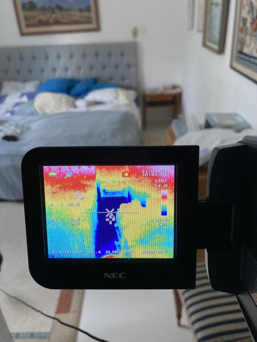 Värmekamerabild som visar en kall punkt i ett rum, kameraskärmen framför suddig interiör.
