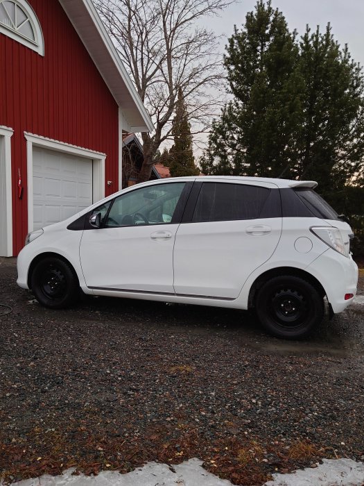 Vit Volvo parkerad på grusuppfart med rentvättade fälgar framför rött hus och garage.