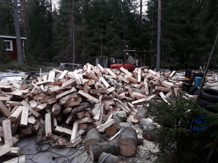 Hög med nysågad klyvd ved och skogsmiljö med byggd vedklyv i bakgrunden.