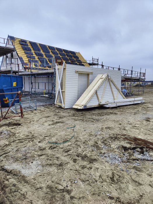 Byggarbetsplats med halvfärdigt hus och takstolar på marken, byggmaterial och ställningar i bakgrunden.