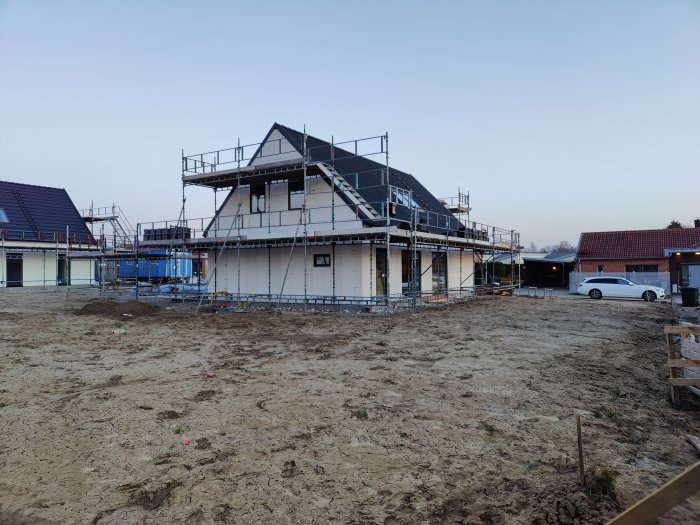 Byggarbetsplats med ett tvåvåningshus under uppbyggnad, byggnadsställningar runt huset och byggmaterial på marken i skymningen.