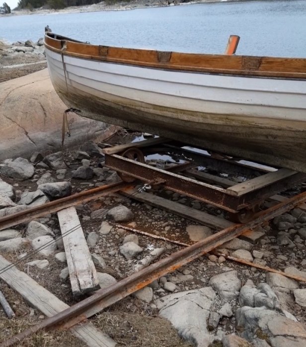 Båt placerad på en gammal rälsbaserad båtupptagningsanordning vid en stenig strandkant.