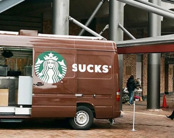 Brun skåpbil med logotyp, texten "SUCKS" syns till höger om logotypen, parkerad vid byggnad.