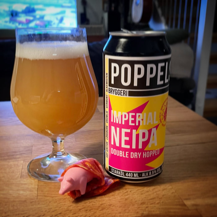 Ölglas fyllt med disig dryck bredvid en burk Poppels Imperial NEIPA och en gummigris insvept i skinka.