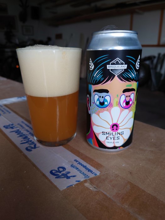 Ölglas med skum vid sidan av en färgglad burk öl märkt "SMILING EYES Triple IPA" på ett brunt bord.