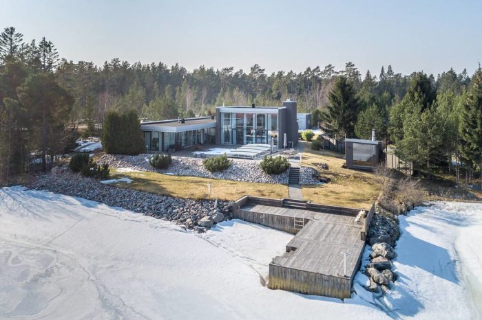 Modern villa med stora fönsterpartier och flat taklinje belägen vid en snötäckt strandkant med skog i bakgrunden.