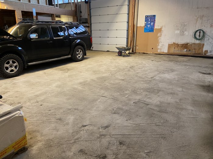 Ett garage med ojämnt betonggolv och en pickup truck, skottkärra och byggmaterial i förgrunden.