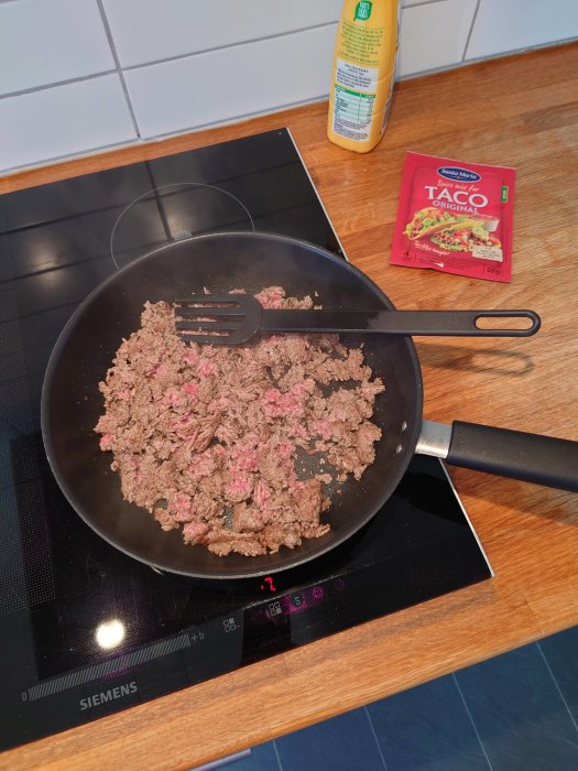 Stekpanna med stekt köttfärs på spishäll, taco-kryddpaket och matoljeflaska i bakgrunden.