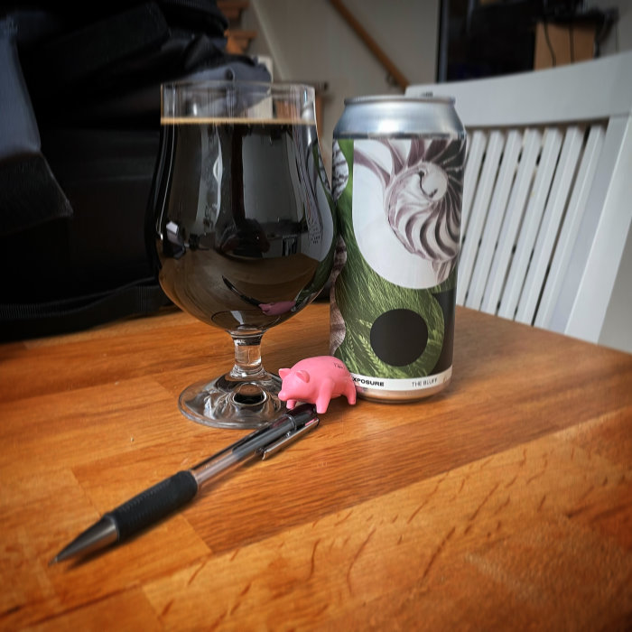 Ölstil i glas bredvid en ölburk och en liten rosa grisfigur ligger på ett träbord med suddig bakgrund.