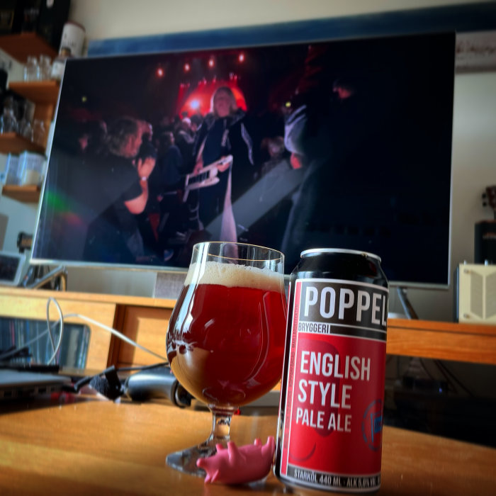 Ölglas och burk från Poppels bryggeri på bord framför en TV som visar en konsert.