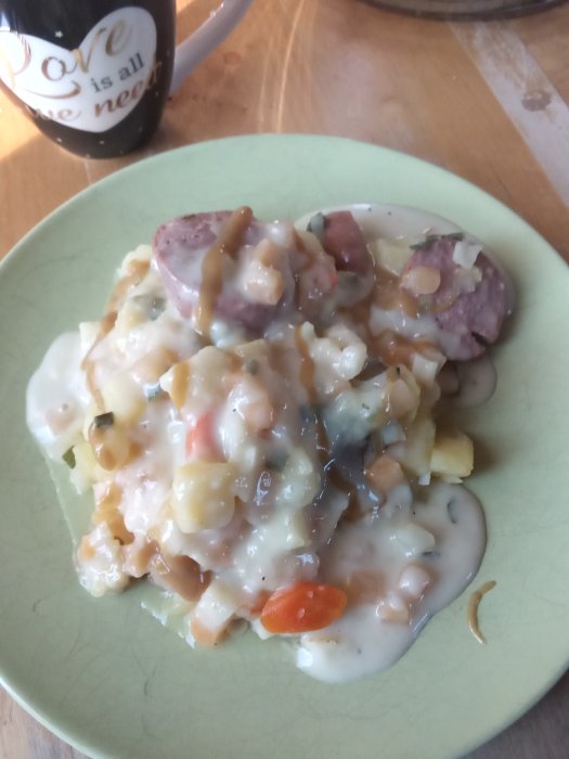 Tallrik med hemlagad rotfruktsgryta och korv täckt av persiljesås, pepparrot tillagd, kopp i bakgrunden.