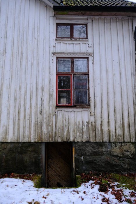 En äldre vit träfasad med rödmarkerade fönster ovanför en öppen källaröppning i en stensockel med snö framför.