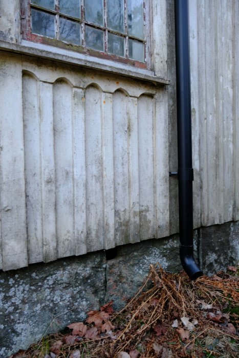 Nedsliten husgrund med äldre stenarbeten sett vid en byggnads hörn med ett nedrör och löv på marken.