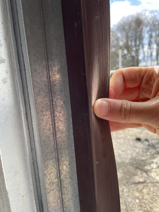 Hand håller en brun plastlist vid en garageport, markerat för att visa skicket.
