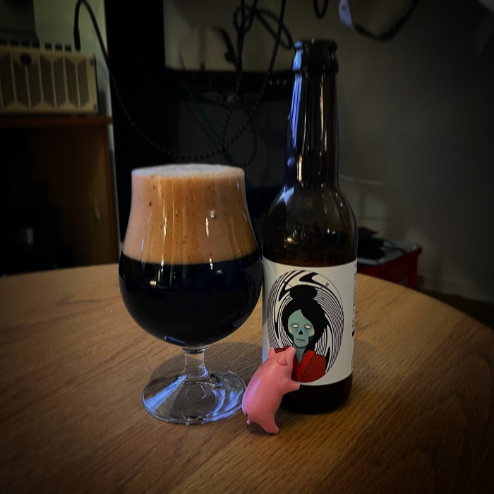 Mörkt öl i glas med tjockt skum bredvid en ölflaska med etikett som föreställer en karaktär.