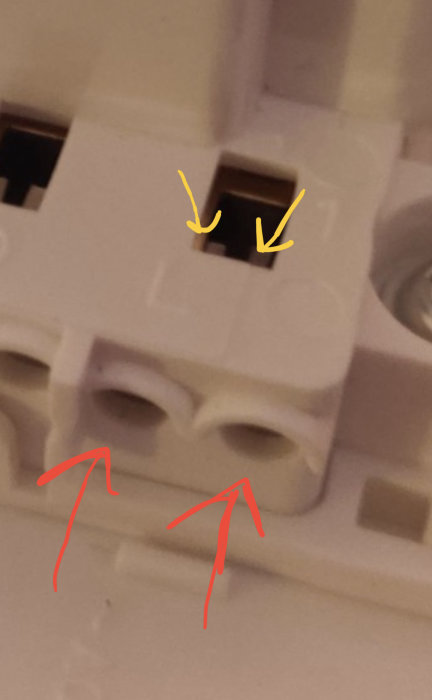 Elektrisk kopplingsklem med runda hål för trådar och metallflärp markerade med röd och gul pil.