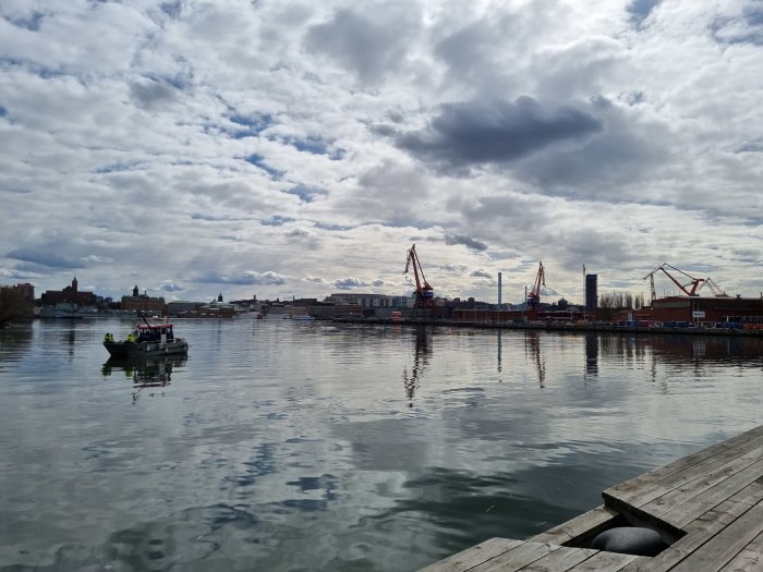 Ett arbetsfartyg på lugnt vatten med stadssilhuett och industrihamn i bakgrunden, delvis molnig himmel ovanför.