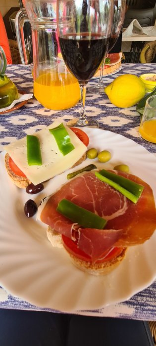 Brödskivor toppade med ost, tomat och prosciutto, oliver och paprika, ett glas rödvin och juicekanna i bakgrunden.