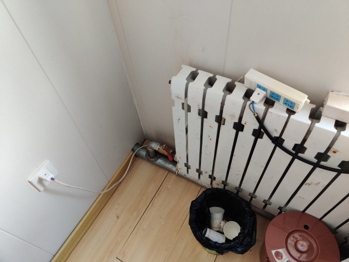 Väggmonterad radiator med avflagad färg och en papperskorg full av skräp i ett hörn av rummet.