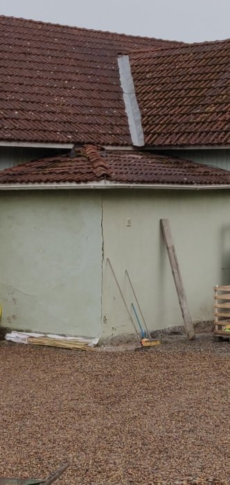 Utsidan av ett hus med granitgrund, sliten puts, tegeltak och trädgårdsredskap vid väggen.