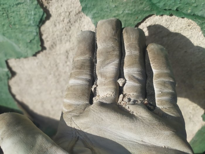 Hand i arbetsvante visar smulig puts från en husgrund med skalad färg i bakgrunden.