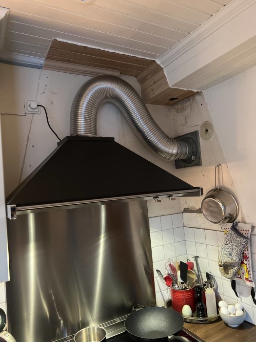 Kök med fläkt och synligt rör som går åt höger, oavslutade väggytor och köksredskap.