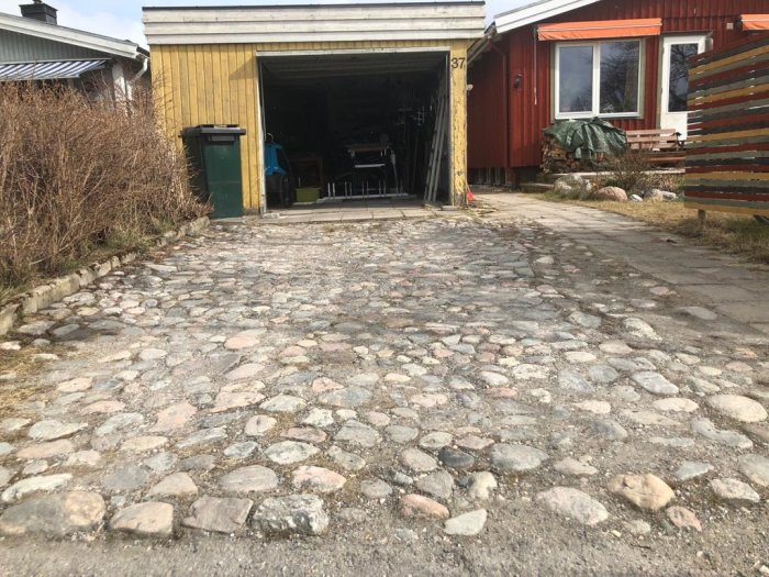 Stenlagd uppfart leder till ett öppet garage, intill ett hus med gult och rött träpanel, synliga sättningar i stenläggningen.