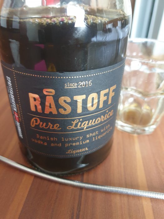 Flaska med RASTOFF Pure Liquorice, dansk lyxshot av vodka och lakritslikör, på ett bord med isfylld shotglas i bakgrunden.