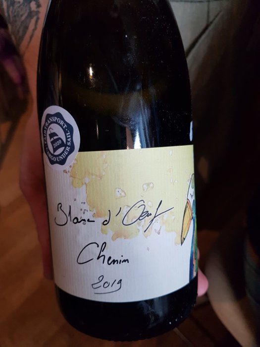 Hand håller en flaska naturvin med etikett "Soirée d'Octobre Chenin 2019" och spillmärken.